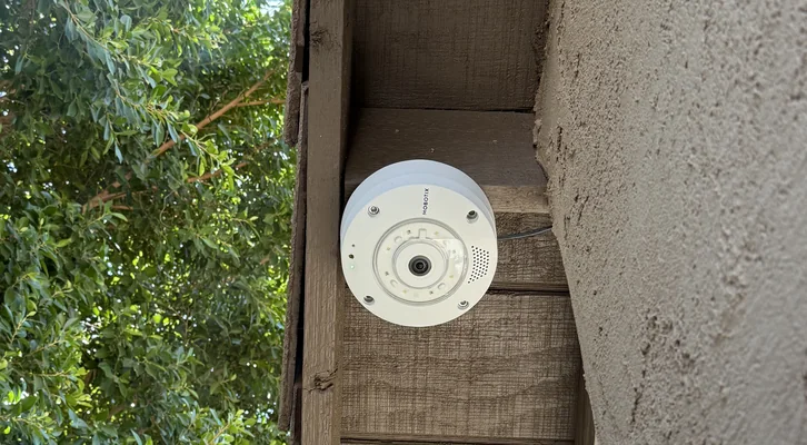 A MOBOTIX Q71 fisheye camera mounted in a timber housing under a concrete soffit, greenery behind it